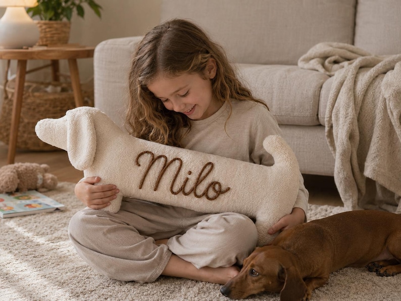 May include: A cream-colored, plush, dachshund-shaped pillow featuring the name Milo embroidered in brown script lettering on its side. The pillow has a soft, textured fabric surface, a long body, floppy ears, and a small tail.