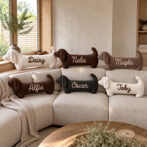 May include: Six plush dachshund-shaped pillows in white, dark brown, and charcoal gray. Each pillow features embroidered names in cursive script: Daisy, Nala, Maple, Alfie, Oscar, and Toby. The pillows have a soft, textured fabric finish.