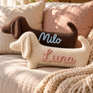 May include: Two dachshund-shaped throw pillows made of textured, curly fabric. The dark brown pillow features the name Milo in light blue script, while the cream-colored pillow features the name Luna in pink script, both stitched onto the side.