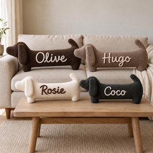 May include: Four plush dachshund-shaped pillows made of textured sherpa fabric. Two pillows are on a sofa, one dark brown with the name Olive and one light brown with the name Hugo. Two are on a table, one cream with the name Rosie and one charcoal with the name Coco.