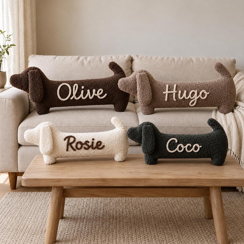 May include: Four plush dachshund-shaped pillows made of textured sherpa fabric. Two pillows are on a sofa, one dark brown with the name Olive and one light brown with the name Hugo. Two are on a table, one cream with the name Rosie and one charcoal with the name Coco.