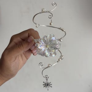 May include: A silver-toned wire ear cuff featuring a spiralling design adorned with small white beads and a central iridescent flower-shaped ornament. A small, dangling star-shaped charm hangs from the bottom of the wire structure.