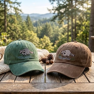 May include: Two baseball caps, one in sage green and one in brown, each featuring an embroidered graphic of a raccoon standing in water. The caps have a structured crown, a curved brim, and eyelets for ventilation, displayed on a wooden surface.