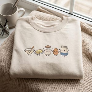 May include: A beige crewneck sweatshirt featuring an embroidered graphic of five anthropomorphic baking ingredients: a bowl with a spoon, a stick of butter, a jar of flour, an egg, and a carton of milk, all with smiling faces and thin limbs.