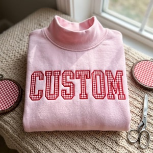 以下が含まれることがあります： 赤と白のギンガムチェック柄で「CUSTOM」という文字が刺繍された、ライトピンクのモックネックトレーナー。ブロック体の文字が胸元に縫い付けられており、長袖で首元が高い丸襟のデザインになっています。