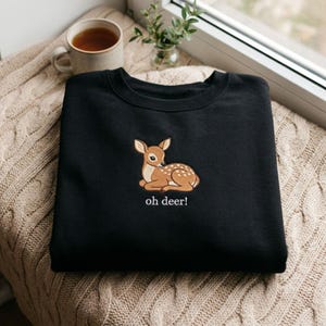 Könnte beinhalten: Ein schwarzes Sweatshirt mit Rundhalsausschnitt und einer Stickerei eines liegenden braunen Rehkitzes mit weißen Punkten. Unter dem Rehkitz ist der Text "oh deer!" in Weiß gestickt. Das Sweatshirt hat lange Ärmel und einen gerippten Kragen.