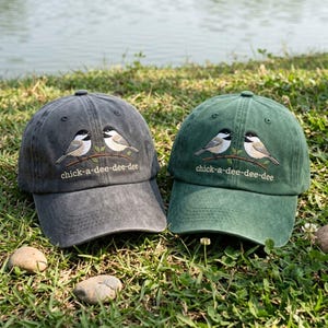 Puede incluir: Una gorra de béisbol gris carbón y una gorra de béisbol verde bosque, cada una con un diseño bordado de dos carboneros posados en una rama con el texto chick-a-dee-dee-dee cosido en hilo blanco debajo de las aves.