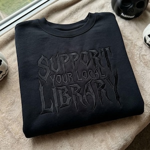 Puede incluir: Una sudadera negra de cuello redondo con bordado negro en el pecho que dice SUPPORT YOUR LOCAL LIBRARY en una fuente gótica estilizada y dentada. La prenda tiene un cuello acanalado y mangas largas, presentada en una vista plana.