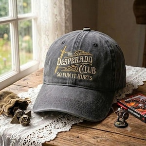 May include: A charcoal grey baseball cap featuring an embroidered gold design with a sword graphic and the text The Desperado Club So Fun It Hurts. The cap has a structured crown, a curved brim, and a distressed fabric finish with visible stitching.