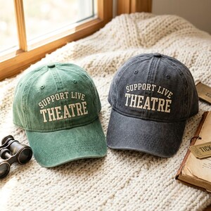 May include: Two baseball caps, one in sage green and one in charcoal gray, both featuring the embroidered text "SUPPORT LIVE THEATRE" in a cream-colored serif font. Each cap has a structured crown, a curved brim, and a distressed fabric finish.