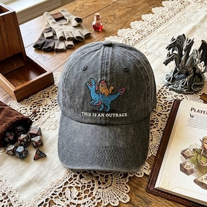 May include: A charcoal grey baseball cap featuring an embroidered graphic of a blue dinosaur with a small orange creature on its back, wearing a pink crown. Below the graphic, the text reads, "THIS IS AN OUTRAGE" in white capital letters.