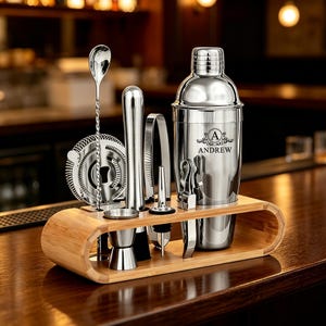 May include: A stainless steel cocktail shaker set with a bamboo stand. The shaker features the engraved letter A and the name ANDREW. The set includes a mixing spoon, a strainer, a jigger, a muddler, a pourer, and tongs, all organized in the stand.