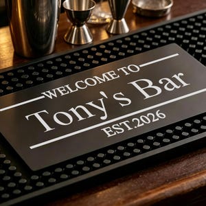 Può includere: Un tappetino da bar rettangolare in gomma nera con una superficie testurizzata a piccoli rilievi. Al centro è presente una scritta bianca che recita "WELCOME TO Tony's Bar" con "EST.2026" sotto, incorniciata da due linee orizzontali bianche.