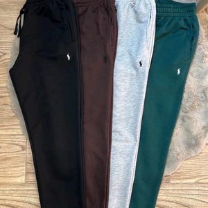 Op de afbeelding: Vier joggingbroeken in zwart, donkerbruin, gemêleerd grijs en donkergroen. Elk paar heeft een elastische tailleband met trekkoord, zijzakken en een klein geborduurd Polo-logo op de linkerpijp, met een blauw Polo-label eraan bevestigd.