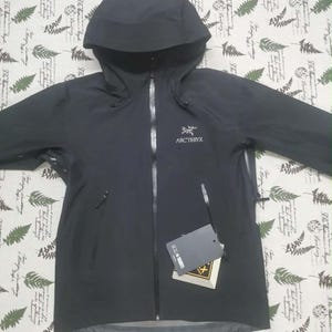 Puede incluir: Una chaqueta negra con capucha Arc'teryx que cuenta con una cremallera frontal completa, mangas largas y dos bolsillos laterales con cremallera. El logotipo de la marca está bordado en el pecho izquierdo. Una etiqueta cuelga de la cremallera.
