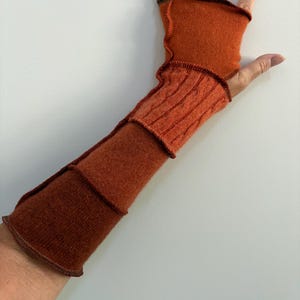 May include: A fingerless glove in a burnt orange hue, featuring a combination of smooth knit fabric and a vertical cable knit pattern. The garment is constructed with visible, dark-toned exposed seams that connect the different textured panels.