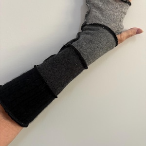May include: A fingerless arm warmer featuring a patchwork design of black, charcoal gray, and light gray knit fabric panels. The garment is constructed with visible black overlock stitched seams and a ribbed texture on the black section.