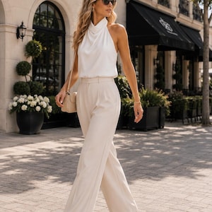 May include: A white sleeveless halter top with a draped neckline paired with high-waisted beige wide-leg trousers. The outfit includes a small beige shoulder bag and beige strappy high-heeled sandals. The top features a smooth fabric finish.