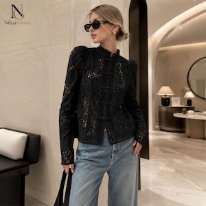 May include: A black long-sleeved blouse featuring intricate lace embroidery, a high collar, and a front button closure with decorative loops. The blouse is paired with light blue denim jeans and black sunglasses, creating a structured look.