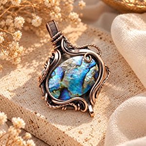 May include: A pendant featuring an oval-shaped abalone shell with iridescent blue and green tones, encased in a dark, oxidised copper wire-wrapped frame. The metalwork includes intricate twisted wire details and a secure bail at the top.