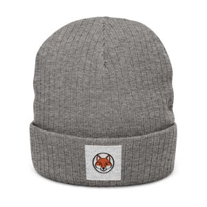 May include: A heather gray ribbed knit beanie featuring a folded cuff. Centered on the cuff is a square white patch displaying a circular black outline containing an embroidered orange and white fox face with black eyes, nose, and ear details.