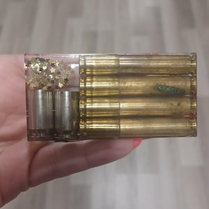 May include: A rectangular clear resin block containing four horizontal brass bullet casings on the right side, and on the left side, two silver-colored metal casings positioned below a cluster of small, gold-colored star-shaped sequins.