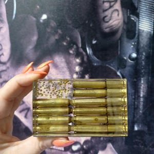 May include: A rectangular clear resin block featuring embedded brass-colored bullet casings arranged in horizontal rows, with a section of small gold star-shaped glitter accents integrated into the transparent material.