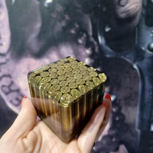 May include: A square-shaped block made of clear resin containing a grid of brass-colored bullet casings. The top surface displays the circular bases of the casings, while the sides reveal the vertical length of the metallic cartridges embedded within.