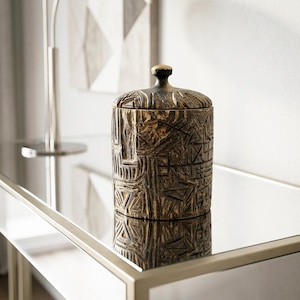 Peut inclure: Un récipient cylindrique en bois avec un couvercle amovible, présentant une finition brun foncé et des motifs géométriques profondément sculptés sur toute sa surface. Le couvercle est surmonté d'un petit bouton arrondi, grain visible.