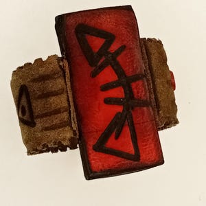 Peut inclure: Une bague avec une pièce centrale rectangulaire rouge ornée d'un symbole noir dessiné à la main. L'anneau est composé d'une matière texturée marron semblable à du daim, avec un petit triangle noir visible sur le côté, au design rustique.