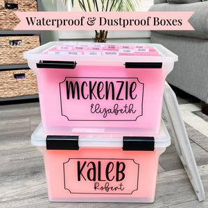 May include: Two stackable, clear plastic storage bins with black locking latches. The top bin contains pink hanging file folders with labels. The front of each bin features a black vinyl decal with names: Mckenzie Elizabeth and Kaleb Robert.