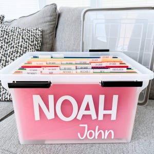 May include: A clear plastic storage bin with a lid, containing colorful hanging file folders labeled with school grades from preschool to fifth grade. The front of the bin features the white vinyl text NOAH in large letters and John below it.