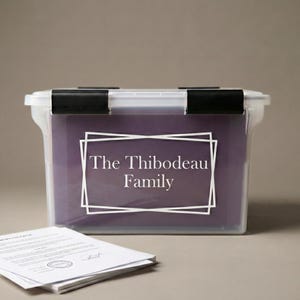 May include: A clear plastic storage bin with two black locking latches on the lid. A purple insert inside the bin features the white text The Thibodeau Family framed by two overlapping white rectangular outlines. A stack of white papers sits in front.