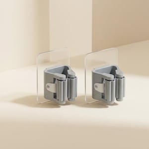 May include: Two gray wall-mounted mop and broom holders featuring a clear adhesive backing plate. Each holder consists of a sturdy gray plastic housing with a spring-loaded roller mechanism designed to grip handles securely in place.