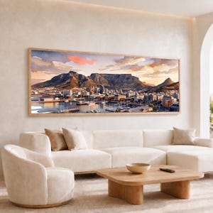 May include: A panoramic landscape painting in a light wood frame depicting Table Mountain overlooking a city harbor with boats at sunset. The sky features soft pink and orange clouds above the dark mountain range and the urban waterfront area.