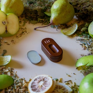 May include: A dark brown wooden oval container with the word "tir" printed in white on the lid, featuring a recessed centre. Beside it lies a small, smooth, light green oval solid balm. The items are surrounded by green apples, pears, and citrus.
