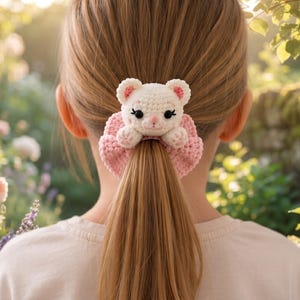 May include: A crochet hair scrunchie featuring a small, white, stuffed animal-like cat head with pink inner ears and black eyes, attached to a ruffled, light pink, textured fabric band that secures a ponytail of straight, light brown hair.
