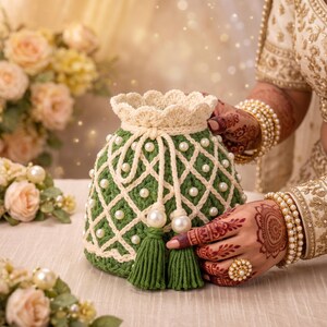 May include: A green crochet drawstring pouch featuring a white lattice overlay adorned with white faux pearls. The bag has a scalloped top edge and two green tassels hanging from the drawstring cords, which are finished with large white pearl beads.