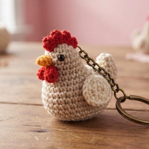 May include: A beige crochet chicken keychain featuring a red comb and wattle, a yellow beak, and black bead eyes. The small stuffed figure is attached to a bronze-toned metal chain and a circular split ring for keys, showcasing detailed yarn stitching.