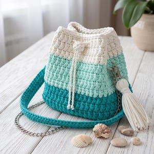 May include: A crochet bucket bag featuring a color-blocked design in white, light mint green, and dark teal. The bag includes a white drawstring closure, a matching teal crochet shoulder strap with silver-tone metal chain links, and a white tassel.