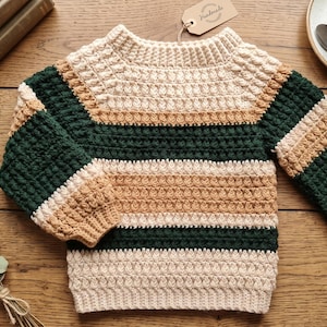 Op de afbeelding: Een gehaakte trui met lange mouwen en horizontale strepen in crème, beige en donkergroen. Het kledingstuk heeft een gestructureerd, bobbelachtig steekpatroon, een geribbelde ronde hals en geribbelde boorden aan het uiteinde van elke mouw.