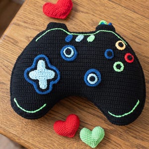 May include: A black crocheted video game controller pillow featuring a light blue directional pad, various colored circular buttons, and neon green accents. The controller is surrounded by small red and light green crocheted heart shapes.