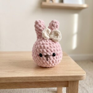 May include: A small, round, light pink crocheted plush rabbit with two upright ears. The rabbit features a cream-colored crocheted bow attached to one ear, two small black plastic eyes, and a stitched brown nose and mouth detail.