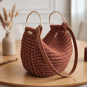 May include: A crescent-shaped handbag made of rust-colored crocheted yarn with a textured bobble stitch pattern. It features two circular gold-tone metal handles and a long, flat tan leather shoulder strap attached with gold-tone metal rivets.