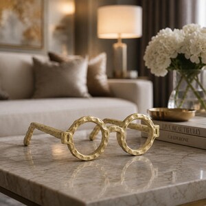 May include: A decorative sculpture shaped like a pair of round eyeglasses with a textured, hammered gold finish. The object features two circular frames connected by a bridge and two side arms, resting on a flat surface.