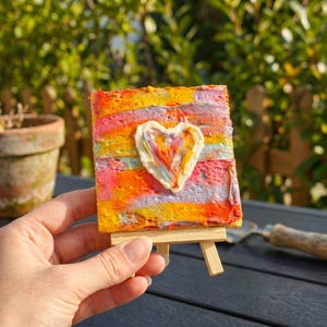 Könnte beinhalten: Ein kleines quadratisches Leinwandgemälde mit einem dicken, strukturierten Impasto-Herz in Weiß, Gelb und Orange in der Mitte. Der Hintergrund besteht aus horizontalen, vermischten Pinselstrichen in leuchtenden Rosa-, Gelb-, Orange- und Hellblautönen.