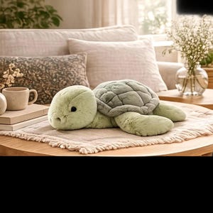 Può includere: Un peluche a forma di tartaruga con morbida pelliccia verde chiaro e un guscio segmentato verde scuro. Il giocattolo ha una testa arrotondata, piccoli occhi scuri e quattro pinne, appoggiato su una superficie in tessuto testurizzato con bordi sfrangiati.
