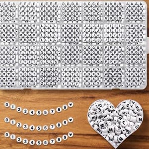 May include: A rectangular clear plastic organizer box filled with white round beads featuring black letters and numbers, alongside three strings of beads displaying numbers zero through nine and heart symbols, plus a heart-shaped container of beads.
