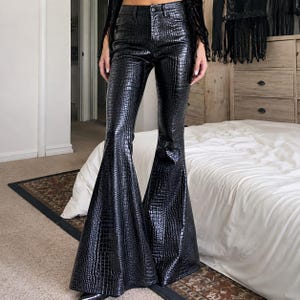 May include: High-waisted black trousers featuring a glossy finish and a textured crocodile skin pattern. These trousers have a fitted waist and hip area that transitions into a dramatic, wide flared leg silhouette, extending down to the floor.
