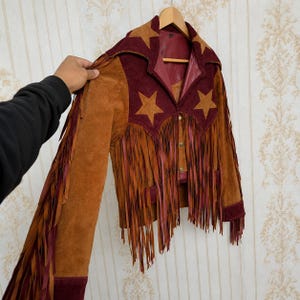 May include: A suede jacket featuring a color-blocked design in tan and deep burgundy. The garment includes star-shaped appliqués on the shoulders and chest, long fringe detailing along the sleeves and front, and a button-down front closure.